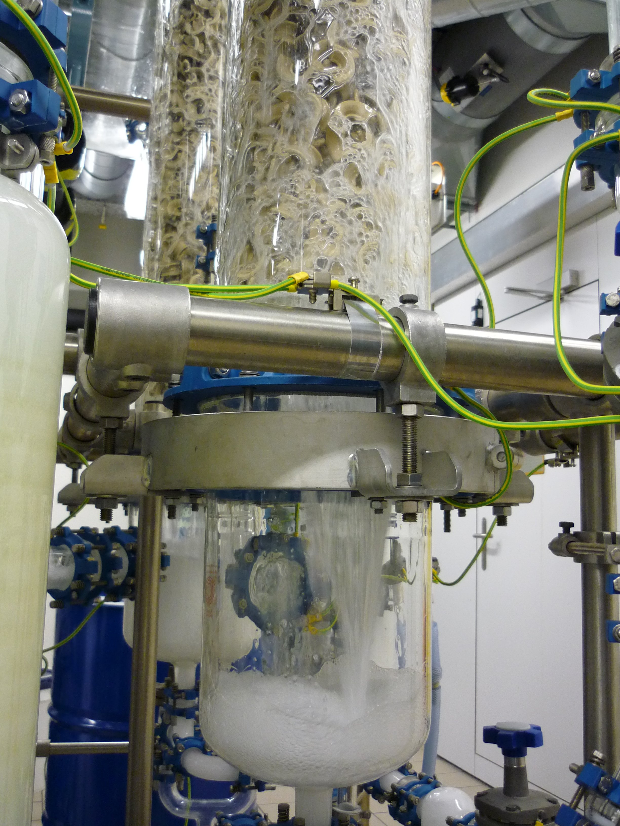 This close up captures the bottom outlet of the glass scrubbing column in operation. The scrubbing liquid passes through the packed bed and drains into the glass receiver below, forming a light foam. This transparent design allows for direct visual monitoring of the liquid flow rate, draining behavior, and phase separation at the column base.